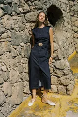 Vestido midi de denim azul oscuro, con cuello redondo y sin mangas. Presenta detalle de frunces en la cintura, cinturón elástico con hebilla dorada y cierre lateral con cremallera oculta. Tiene falsos botones delanteros.