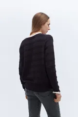 Sweater de punto negro con detalle de calados y lazo en el escote. Tiene ribetes con volados en contraste en el cuello y puños.