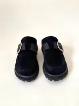 Zueco tipo mule negro, confeccionado en cuero vacuno, con puntera cuadrada y suela track gruesa. Presenta una faja ancha con hebilla metálica plateada sobre el empeine.