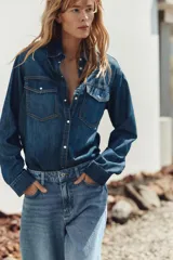 Camisa de denim azul con cuello solapa, manga larga, dos bolsillos delanteros con solapa y cierre frontal con botones a presión.