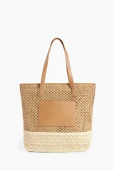Bolso tipo tote de raffia color tostado, con base tejida en color crema y aplique rectangular de cuero sintético en el frente. Tiene doble asa de hombro de cuero sintético color camel.