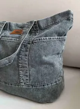 Bolso grande tipo totebag de jean azul, con asas de mano y asa larga para colgar. Se cierra mediante 1 botón de metal a presión y contiene 3 bolsillos exteriores.