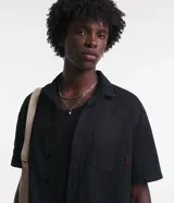 Camisa negra de sarga, corte boxy, con cuello inglés, manga corta, abotonadura frontal y bolsillo en el pecho.