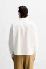 Camisa blanca de corte relaxed fit confeccionada en tejido de algodón y lino. Tiene cuello bowling, manga larga con puños abotonados, jaretas en el delantero, bolsillos de parche en pecho y cadera, bajo con aberturas laterales y cierre frontal con botones.