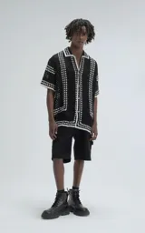 Camisa de manga corta color negro, tejida en punto abierto (símil crochet) con detalles en blanco. Tiene cuello tipo solapa, botones frontales y un calce oversize.