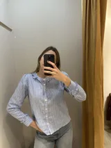 Camisa celeste a rayas blancas, de algodón y lino, con cuello mao y volados en cuello y mangas. Cierre frontal con botones blancos.