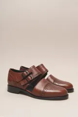 Zapato bajo de cuero negro, estilo monk strap con dos hebillas ajustables sobre el empeine. Presenta detalles de brogue (perforaciones) en la puntera y alrededor del borde superior. Tiene un taco bajo y ancho.