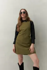 Vestido corto de algodón color verde oliva, de corte recto, con cuello redondo y apliques metálicos en el escote y sisas. Se lleva sobre una camisa negra de manga larga.