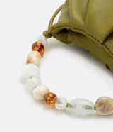 Bolso pequeño color verde oliva, desestructurado, con pinzas frontales y cierre de botón a presión. Incluye una correa de hombro extraíble y un asa de mano decorada con cuentas acrílicas variadas en tonos blancos, marrones y transparentes.