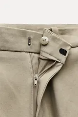 Pantalón de vestir color beige, de tiro alto y corte recto. Incluye cinturón marrón con hebilla metálica. Presenta bolsillos laterales y falsos bolsillos traseros de doble vivo. Cierre frontal con cremallera, botón interior y gancho oculto.