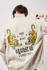 Buzo color arena de corte oversized, con cuello redondo y puños y cintura acanalados. Estampado en la espalda con dos tigres enfrentados y texto "Pray x Mercy" y "Against All Since XIX".