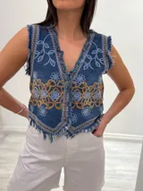 Chaleco corto de denim azul con bordados florales en blanco y marrón, detalle desflecado en escote, sisas y ruedo. Escote en V y cierre frontal con botones. Estilo boho chic.