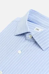 Camisa celeste a rayas verticales blancas, de corte recto, con cuello italiano abierto, manga larga con puños abotonados y cierre frontal con botones.