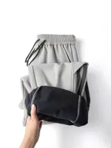 Pantalón deportivo gris claro, estilo cargo o jogging, con franjas negras verticales en los laterales de las piernas. Posee tobillos regulables y cintura ajustable.