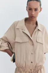 Camisa color beige de cuello solapa y manga corta con trabilla y botón. Tiene bolsillos delanteros de plastrón con solapa, bajo con elástico y cierre frontal con botones a presión.