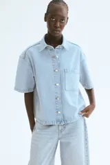 Camisa celeste de denim, corte amplio, con cuello clásico, manga corta, cierre frontal con botones metálicos, bolsillo de parche en el pecho y canesú en la espalda.