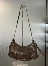 Cartera marrón con textura de cuero, forma de media luna y correa de hombro ajustable. Tiene cierre superior con cremallera y un bolsillo frontal con hebilla decorativa.