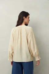 Camisa color crema de lino con cuello redondo, canesú fruncido, mangas largas amplias con puños elastizados con volado y cierre frontal con botones.