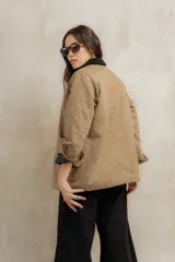 Campera color beige con cuello de corderito negro, cierre con botones, bolsillos laterales y puños con detalle de cuero sintético negro.