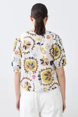 Camisa blanca con estampado floral en tonos azul, amarillo, rosa y marrón. Tiene cuello mao, mangas cortas abullonadas y cierre frontal con botones.