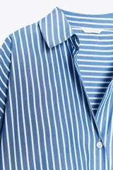 Camisa celeste con rayas verticales blancas, de corte holgado, cuello solapa, escote pico y mangas largas con puños abotonados.
