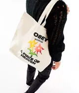 Bolso tipo tote bag de tela color crema con asas largas. Estampado frontal con el logo de Obey, flores y la frase "A Piece of Heaven".