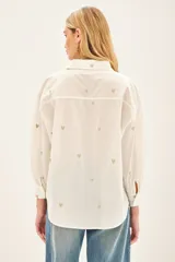 Camisa blanca de algodón con bordado de corazones dorados, corte amplio, cuello clásico, cartera con botones ocultos y bolsillo en el pecho.