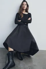 Falda midi negra de gabardina, tiro medio, cintura con trabillas, corte evasé con volumen y cierre frontal con cremallera y botón.