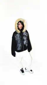 Hoodie negro con cierre, capucha con borde de piel sintética, estampado con texto y cruz con calaveras en el frente.