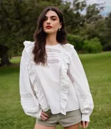 Blusa blanca de tricolina con volados en los laterales, cuello redondo, cierre por gota, mangas abullonadas largas y entremedios en el busto y en las mangas.