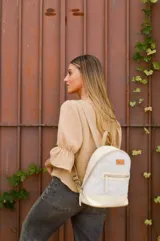 Mochila pequeña de cuero color gris claro y beige, con cierre superior y bolsillo frontal con cierre. Tiene asa de mano y correas ajustables para los hombros.