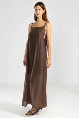 Vestido largo color marrón chocolate, con breteles finos y escote recto. Presenta lazos en los costados, forro hasta arriba de las rodillas y caída posterior más larga.