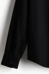 Sobrecamisa negra de corte recto, con cuello clásico, cierre frontal con botones, dos bolsillos de parche con solapa en el pecho, mangas largas con puños abotonados y bajo redondeado.