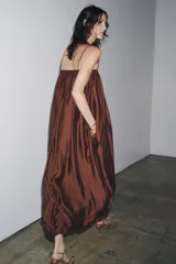 Vestido largo marrón de tejido satinado, con escote recto, tirantes finos extraíbles, bolsillos laterales ocultos y bajo asimétrico acabado en globo.