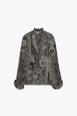 Camisa de viscosa con estampado paisley en blanco y negro, cuello redondo con volantes y escote pico ajustable con cordones. Mangas largas con elástico y volantes en los puños, bajo asimétrico y cierre frontal con botones forrados.
