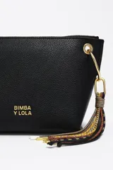 Bolso bandolera de piel vacuna negra, con forma trapezoidal y estructura rígida. Posee asa regulable, cierre de cremallera, bolsillo interior y charm decorativo con borlas en tonos tierra y dorado.