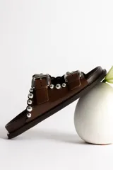 Sandalia tipo mule negra de cuero, con dos tiras ajustables con hebillas metálicas plateadas y apliques de tachas esféricas plateadas. Presenta un borde festoneado en la tira superior y suela de plataforma negra.