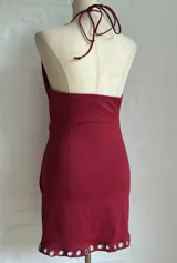 Vestido corto ajustado color rojo oscuro, estilo halter con escote pronunciado en V que se ajusta con ojales metálicos a lo largo del borde. El ruedo también presenta ojales metálicos.