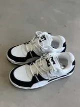 Championes DC Manteca 4, color blanco con detalles en negro. Presentan una parte superior de malla y cuero sintético, con el logo de DC en la lengüeta y el lateral. Cuentan con cordones blancos y una suela de goma.