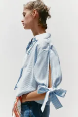 Camisa celeste y blanca a rayas verticales, confeccionada con lino e hilo metalizado. Tiene cuello solapa, mangas 3/4 con lazos en los puños y cierre frontal con botones ocultos por solapa.