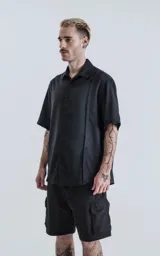 Camisa negra de manga corta con cuello clásico y cierre frontal con botones. Presenta una tira vertical cosida en el frente.