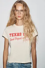 Camiseta color crudo de algodón con efecto lavado, cuello redondo, manga corta con hombro caído y estampado frontal con la leyenda "Texas USA Ranch Original est. 1975" en color rojo.