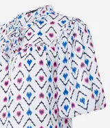Blusa de chiffon con estampado geométrico en tonos blanco, azul y rosa. Tiene cuello redondo con lazo, mangas cortas abullonadas y volados en el cuello y los puños.