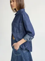 Camisa de denim azul con pespuntes blancos, corte crop y cierre frontal con botones. Tiene cuello clásico, mangas cortas y un bolsillo de parche en el pecho. Se ata en la parte delantera.