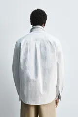 Camisa celeste a rayas verticales blancas, de corte relaxed fit, cuello solapa, manga larga con puños abotonados, bolsillo de parche en el pecho y cierre frontal con botones.