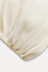 Blusa color crudo confeccionada en hilatura con mezcla de lyocell. Escote pico ajustable con cordones, manga larga con volumen acabada en elástico y detalle de volantes en el mismo tejido.