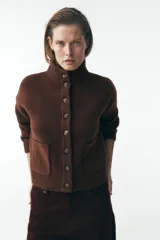 Chaqueta de punto marrón con cuello alto y manga larga. Tiene cierre frontal con botones y bolsillos de parche en el delantero.