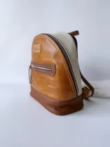 Mochila pequeña de cuero color camel, con base marrón, laterales color crema y cierre superior con cremallera.