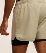 Bermuda deportiva color verde claro con short de compresión interno negro, cintura elástica con cordón ajustable y detalle reflectante trasero.