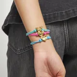 Pulsera ajustable con doble hilo elástico de color celeste brillante, centrada por un dije de corazón tridimensional con acabado dorado. Presenta un cierre o ajuste de forma ovalada dorada en la parte superior y un nudo decorativo a cada lado del corazón.